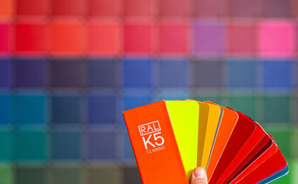 Hand holding a fan of colorful paint swatches in front of a wall covered with a grid of different colors.