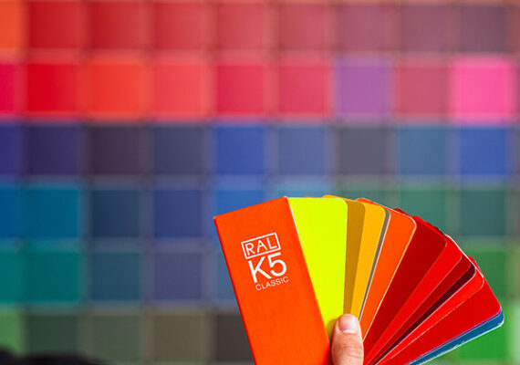 Hand holding a fan of colorful paint swatches in front of a wall covered with a grid of different colors.