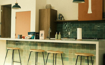 Modern kitchen with green tiled backsplash, wooden bar stools, and minimalist pendant lights over a breakfast counter.