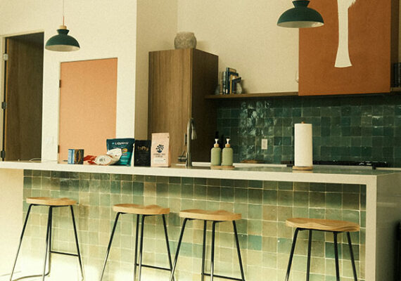 Modern kitchen with green tiled backsplash, wooden bar stools, and minimalist pendant lights over a breakfast counter.