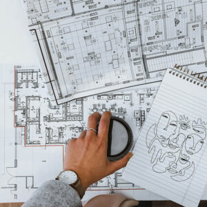 Hand holding a coffee cup over architectural floor plans, with a sketch of abstract faces on a notepad.