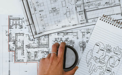 Hand holding a coffee cup over architectural floor plans, with a sketch of abstract faces on a notepad.