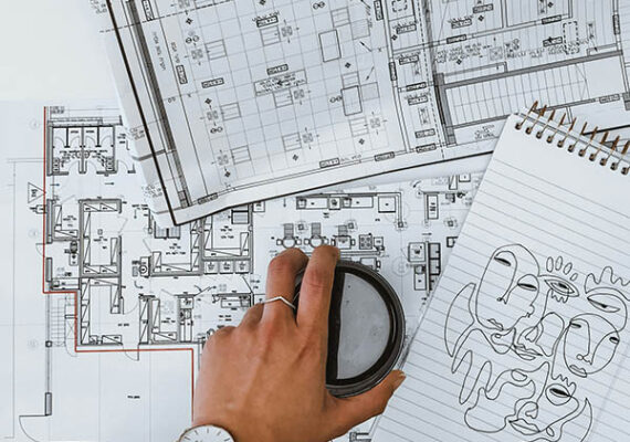 Hand holding a coffee cup over architectural floor plans, with a sketch of abstract faces on a notepad.