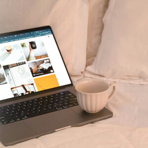 Open laptop on a bed displaying photos, with a white coffee mug resting on the keyboard.