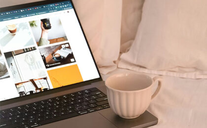 Open laptop on a bed displaying photos, with a white coffee mug resting on the keyboard.