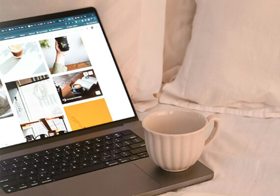 Open laptop on a bed displaying photos, with a white coffee mug resting on the keyboard.